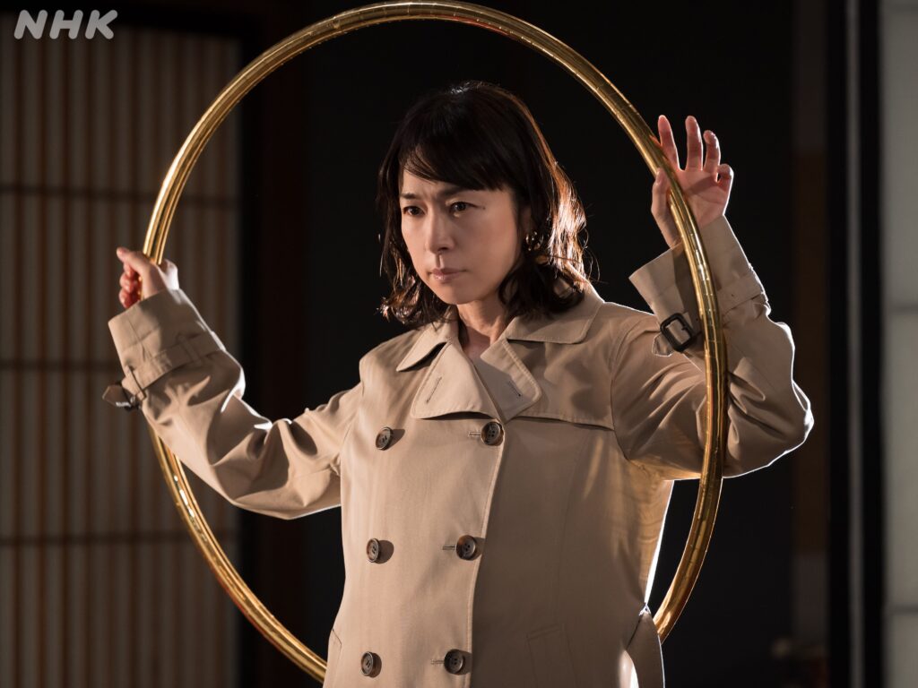NHKのコント番組LIFE!にて女神探偵・輪島ナオミを演じる西田尚美さん。金色のフープを背面に構えている。